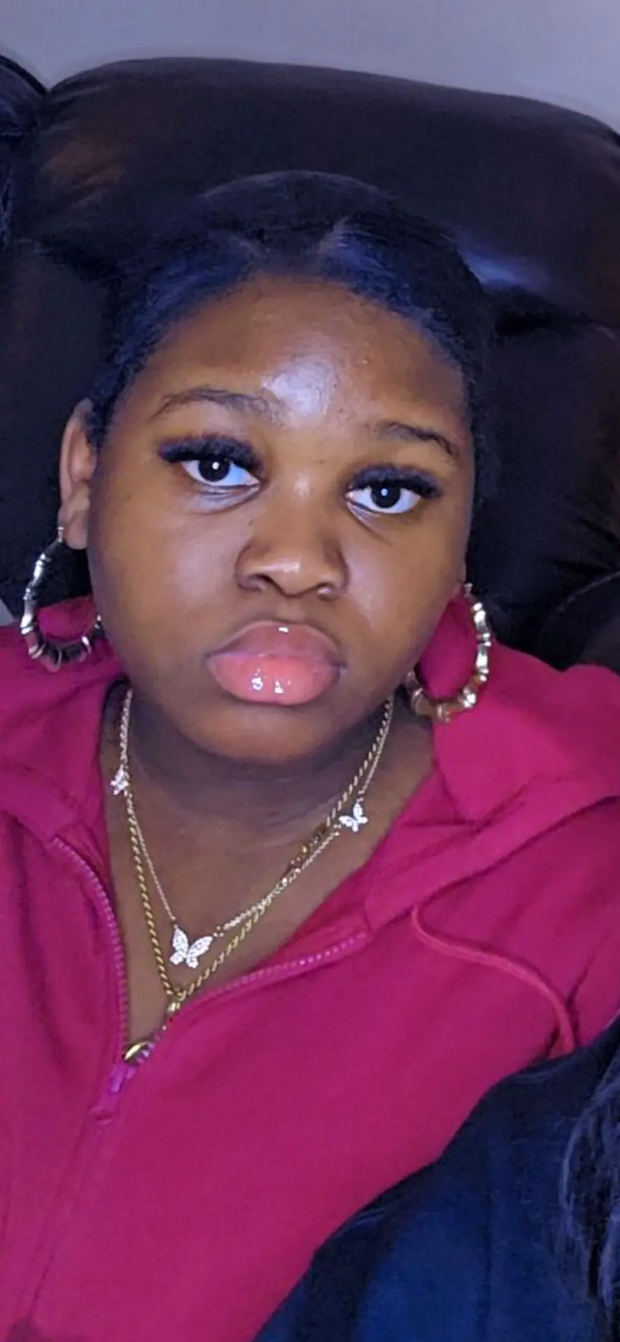 Korkor, founder of Courage Digital Studio, in a close-up portrait: hair parted in the middle and pulled back, wearing a berry-pink zip-up hoodie, gold hoop earrings and layered gold necklaces with butterfly pendants, looking calmly into the camera against a dark leather chair.